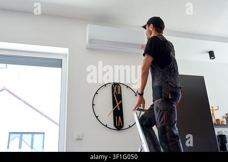 Air conditioner technician stands on a ladder and inspects a wall mounted AC unit in a modern home interior. Man performs maintenance service on the indoor cooling system. Stock Photo