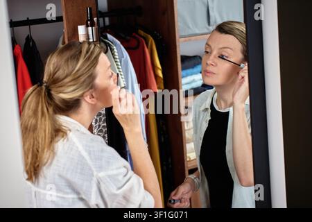 Woman applying mascara while looking in wardrobe mirror, holding a brush near her eye. Concept of beauty routine, makeup and personal care. Stock Photo