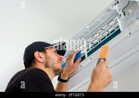 Man cleans a removable filter inside an open wall mounted air conditioner during home maintenance, checks the cooling unit while servicing indoor climate equipment. Stock Photo