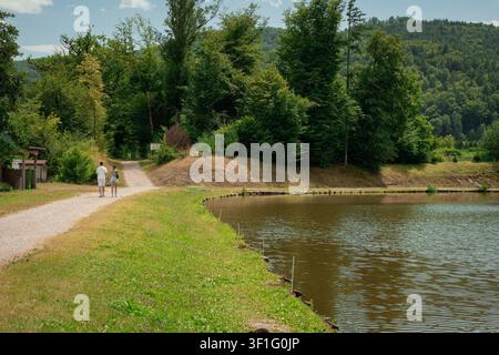 Panoramic view of a tranquil pond near Radece, surrounded by dense green forest and rolling hills, with two people walking along a gravel path in the Stock Photo