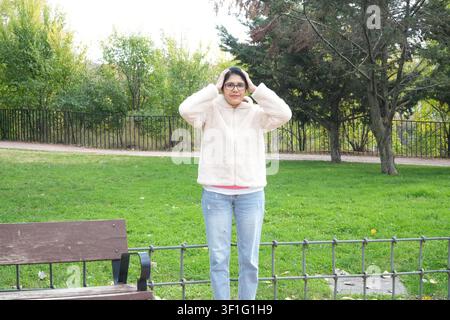 Woman stands outdoors, adjusting her glasses. She wears a fluffy sweater and jeans. Green grass and trees form the background. Bench sits nearby, besi Stock Photo