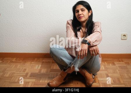Woman sits on floor, knees bent. She wears casual clothing and boots. Her hands rest on her knees. Wall behind her has a textured finish. Floor is woo Stock Photo