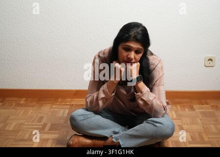 Young woman sits floor, hands clasped under chin. She rests elbows on knees, head bowed. Brown boots and jeans frame her seated pose. Wall behind has Stock Photo