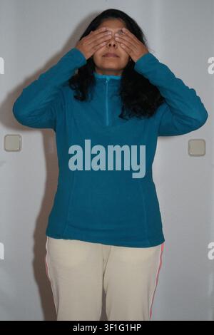 Young woman covers eyes with hands. She wears teal sweater and light pants. Wall behind her has two switches. Her expression is calm and focused. Simp Stock Photo