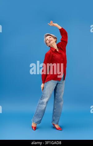 Senior woman with grey hair dances playfully in stylish red blouse and jeans Stock Photo