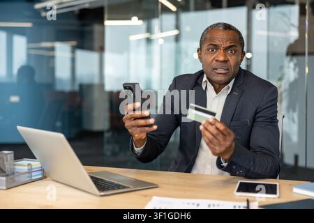 Mature businessman reacting with surprise and confusion, holding a credit card and smartphone while using a laptop, indicating issues with online bank Stock Photo
