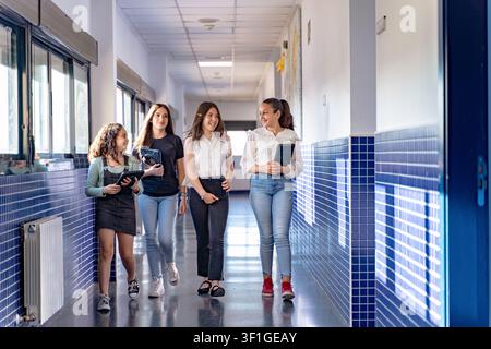 Four happy girls holding digital tablets and files, walking and talking around school corridor during break time Stock Photo