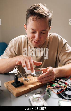 Vertical portrait of technician male examining complex circuit board with magnifying glass, performing detailed electronic repair work, problem Stock Photo