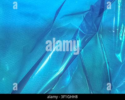 Abstract background featuring textured blue fabric beneath a sheet of wrinkled, reflective plastic. Smooth, shiny surface interplay with matt fabric. Stock Photo