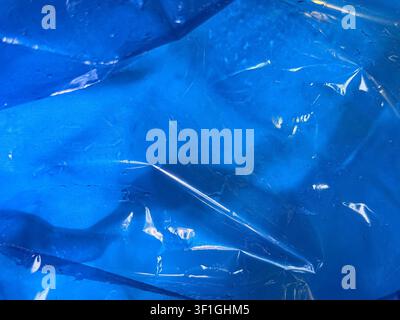 Close up of vivid blue fabric covered by translucent wrinkled plastic sheet. Light sparkles on the surface reflecting from subtle water drops creating Stock Photo