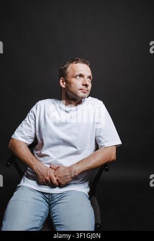 A contemplative casual man sits in a chair wearing a white t-shirt and jeans, gazing upward against a dark studio wall. Clean, minimal portrait convey Stock Photo