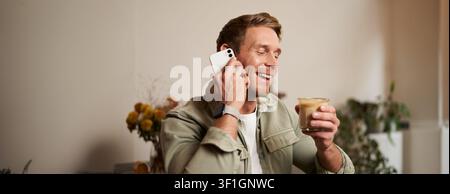 Portrait of young handsome man talking on the phone, drinks coffee and laughs. A guy sits in cafe having a nice, pleasant conversation over cup of cap Stock Photo