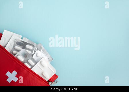Red first aid kit bag opening, revealing blister packs of white medicine pills, capsules, and health supplies, placed on a light blue background Stock Photo