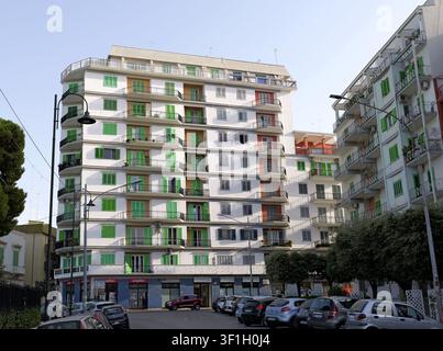 Modern apartment buildings dominating a city street with cars parked below, featuring many windows with green shutters and balconies. Italy, apulia, m Stock Photo