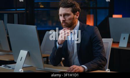 Concentrated Caucasian middle-aged business man working with computer thinking at evening pensive looking in desktop monitor adult mature male Stock Photo