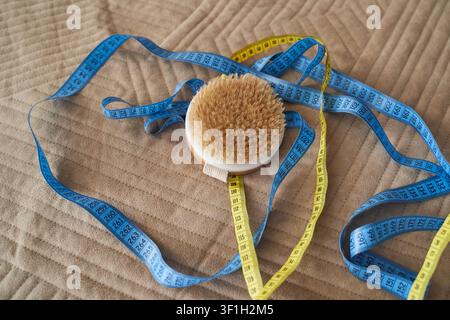 Body Brush Resting Amid Measuring Tapes On Beige Quilted Bedspread, Close-Up Still Life Of Grooming Tool And Flexible Tape Measure In Domestic Setting With Soft Daylight. Stock Photo