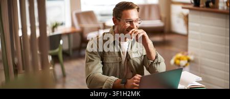 Image of young digital nomad, man in glasses sits in cafe, works from coffee shop, uses laptop in co-working space, wears glasses, drinks his beverage Stock Photo