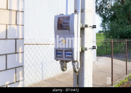 Electrical control box outdoor. Gray plastic distribution panel with circuit breakers mounted on concrete pillar near white brick wall and mesh fence Stock Photo