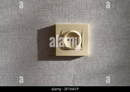 Electric power outlet. Single beige plastic socket installed on grey textured wallpaper wall inside room apartment under side daylight Stock Photo