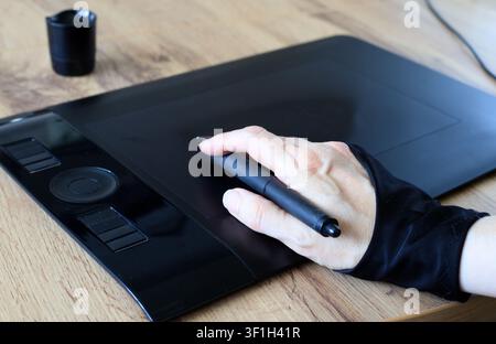Digital artist workspace. Hand in two finger glove holding pen stylus on black drawing tablet over wooden table surface indoor Stock Photo