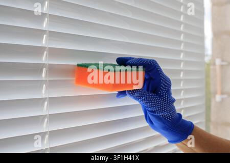 Cleaning venetian blinds texture. Person wiping white plastic slats with abrasive orange and green sponge inside home apartment room under daylight Stock Photo
