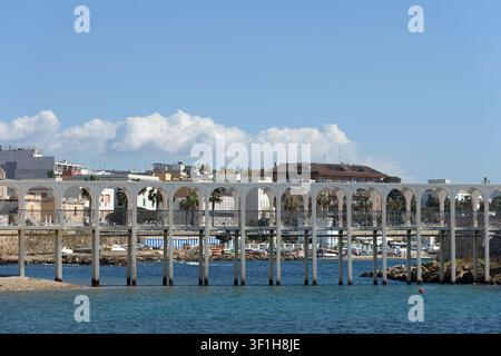 White arched pedestrian bridge over blue sea water with a coastal town and Mediterranean architecture in the background under a sunny sky Stock Photo