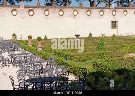 Multiple sets of black wrought iron patio tables and chairs arranged on a gravel terrace in the formal garden of Villa Mondragone, featuring neatly tr Stock Photo