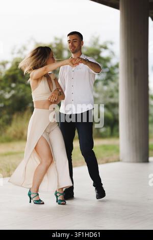 Couple dancing outdoors practicing a romantic Latin partner dance under a pavilion. woman in flowing skirt and heels, man leading in a white shirt Stock Photo