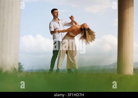 Couple dancing outdoors as man gently dips woman under a sunny sky, romantic partners practicing a graceful dance move near columns Stock Photo