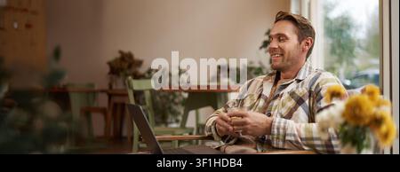 Image of handsome businessman working from cafe, drinking coffee and doing online project on his laptop, smiling and looking happy, finished his freel Stock Photo