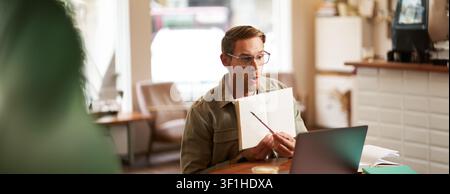 Portrait of handsome young man giving lessons online, showing exercises in his notebook, looking at laptop, recording video, sitting in cafe, quite co Stock Photo