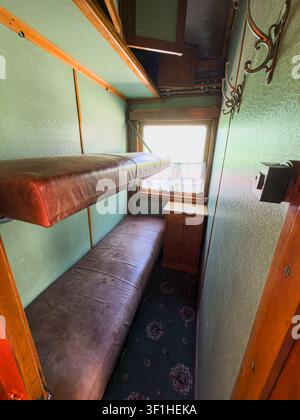 Vintage train car interior featuring luxurious compact bed, wooden nightstand, green walls, leather-like cover, dark carpet, small window, warm natura Stock Photo