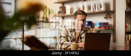 Image of young business owner, man sitting in cafe with laptop, looking confused at screen, digital nomad trying to solve the task, programmer working Stock Photo