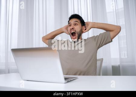 Young shouting man holding head hands with open mouth, experiencing shock and disbelief while looking at up laptop screen, reacting to unexpected bad Stock Photo