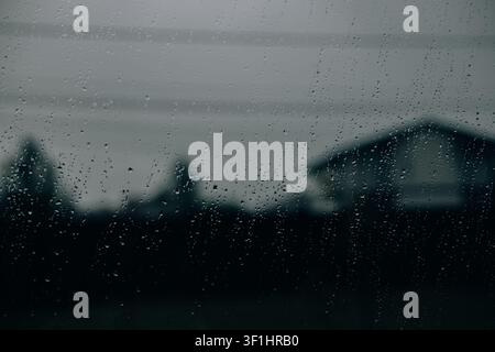 Raindrops on a window during heavy rainfall with blurred residential houses in the background. Atmospheric image representing stormy weather, seasonal Stock Photo