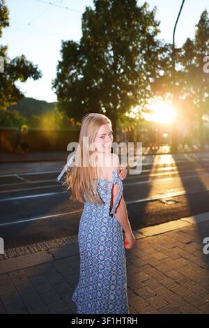 Happy female traveler in a floral blue dress during golden hour, walking through an urban street with warm evening sunshine. Stock Photo
