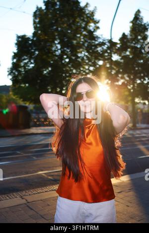 Young brunette enjoying golden hour in the city, wearing sunglasses and trendy clothes, modern fashion and travel concept. Stock Photo