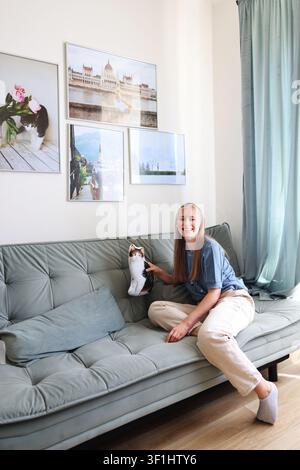 Young woman holding a cat-shaped cushion beside her curious cat on a sofa in a cozy living room. Stock Photo