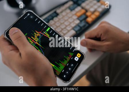 Trader using smartphone analyzing financial market data and stock charts on mobile application, monitoring investments, cryptocurrency prices, and Stock Photo