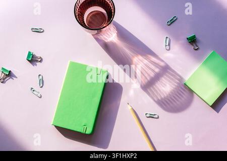 Green notebook with sticky notes, pencil, paper clips and glass on purple background with light and shadows. Top view, flat lay. Stock Photo