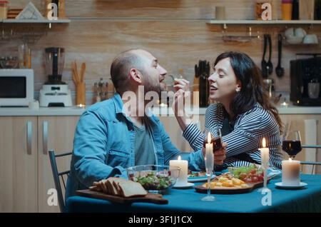 Cheerful couple shares a tender moment during a romantic dinner in the kitchen, dining together and celebrating an anniversary, inside a realistic lifestyle setting. Stock Photo