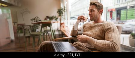 Working people and public space concept. Young handsome man, ux ui designer sitting with laptop in cafe, drinking cappuccino, looking at screen with s Stock Photo