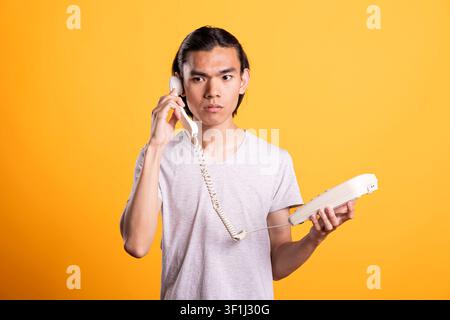 Asian man speaking on phone with concentrated and serious face, reacting to potentially urgent information. Male model holds telephone receiver to his ear listening carefully during phone conversation Stock Photo