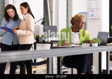 Black corporate employee analyzes project updates on laptop, reviewing documents and maintaining smooth business workflow. African american woman working in productive multiethnic workplace. Stock Photo