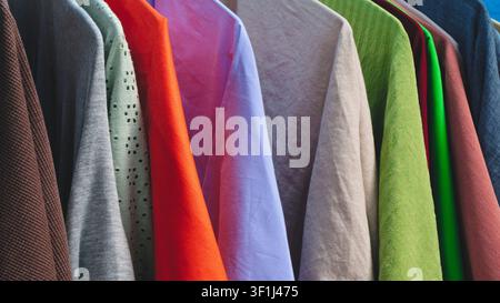 Colorful fabrics hang in a row on a clothing rack. Hues include brown, gray, orange, purple, green, and blue. Each textile features different textures Stock Photo
