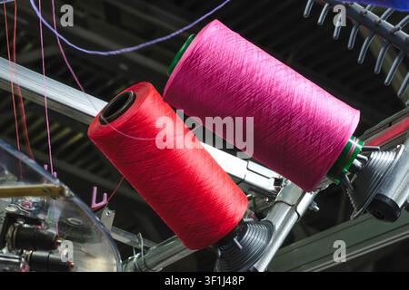Two large spools of thread spin on machine frame. One spool is bright pink,  other is vivid red. Metallic parts and wires surround spools Stock Photo