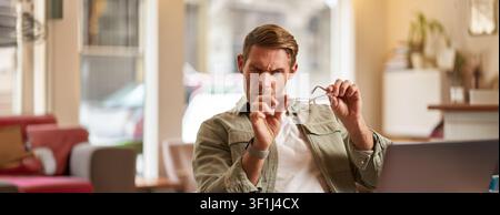 Portrait of man with new anti blue light glasses, looking through the lense at his computer screen, sitting in cafe with laptop and testing eyewear. Stock Photo