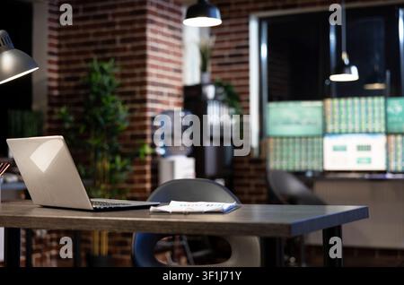 Vacant modern office desk holds a laptop and clipboard against a brick wall at night, with blurred monitors creating a minimalist workspace. Stock Photo
