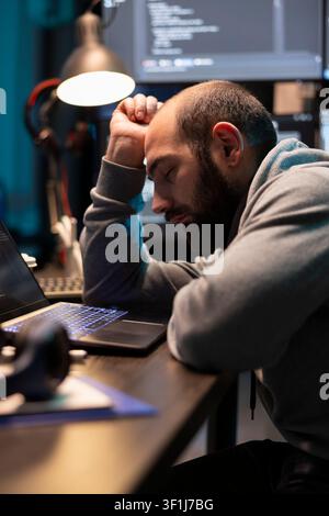 Adult male developer sleeps at desk, feeling tired after working late hours on cybersecurity tasks. Stressed man resting on arm, exhausted from planning and debugging encrypted programs. Stock Photo