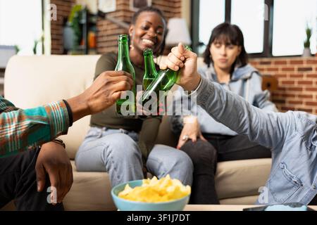 Multiethnic friends sit together on couch, clinking beer bottles in celebration during casual hangout at home. Cheerful diverse group enjoys drinks, snacks and laughter in cozy brick wall apartment. Stock Photo